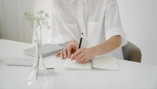 An anonymous person in a crisp white shirt taking notes at a minimalist desk — illustrating Fragrenza's edit of the most polished office-appropriate fragrances.